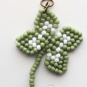 May include: A green and white beaded four-leaf clover charm with a gold jump ring. The charm is handmade and has a delicate, intricate design.