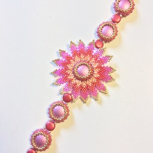 Op de afbeelding: Een roze en oranje kralenarmband met een grote bloemvormige bedel in het midden. De armband is gemaakt van kleine kralen en heeft een gouden sluiting.