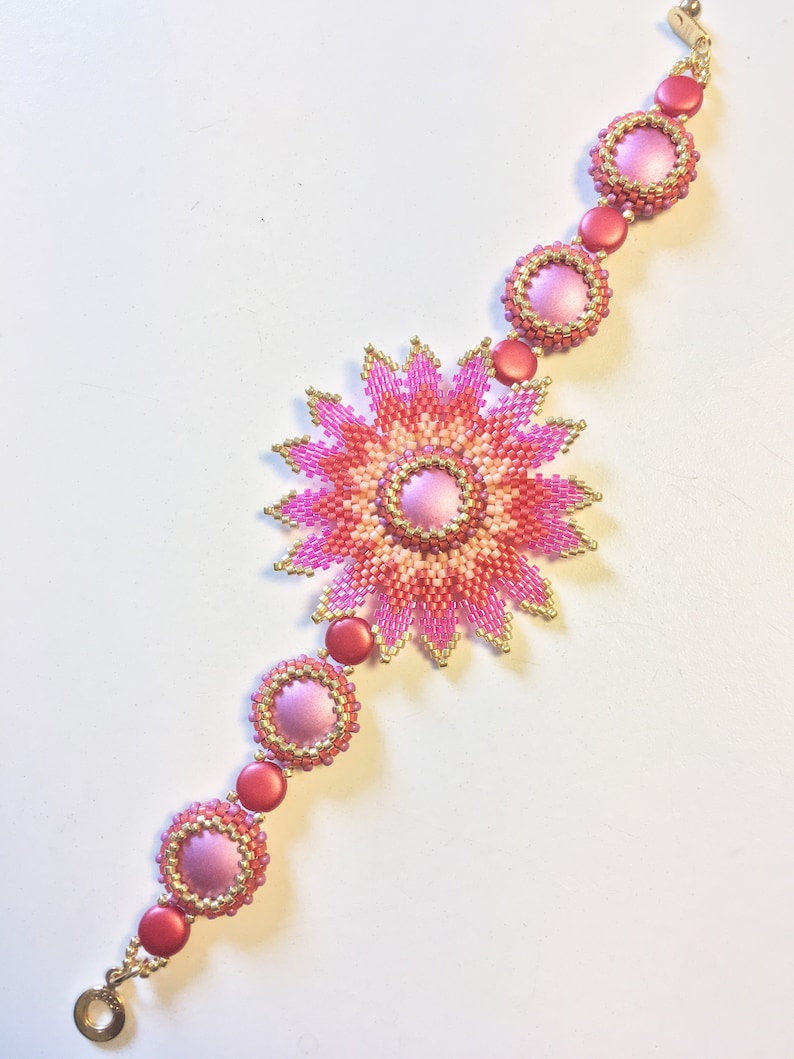 Op de afbeelding: Een roze en oranje kralenarmband met een grote bloemvormige bedel in het midden. De armband is gemaakt van kleine kralen en heeft een gouden sluiting.