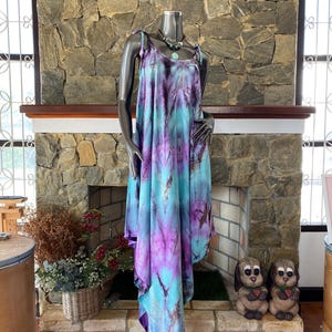 Op de afbeelding: Een tie-dye jurk in paarse en turquoise tinten op een mannequin. De jurk heeft een vloeiend, asymmetrisch ontwerp met spaghettibandjes. Een ketting met een grote hanger wordt gedragen. De achtergrond omvat een stenen open haard en decoratieve elementen.