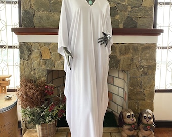 May include: A white, flowing caftan dress with a V-neck and wide sleeves is displayed on a mannequin. The dress is floor-length and appears to be made of a lightweight fabric. A turquoise necklace adorns the mannequin's neck. The background features a stone fireplace.