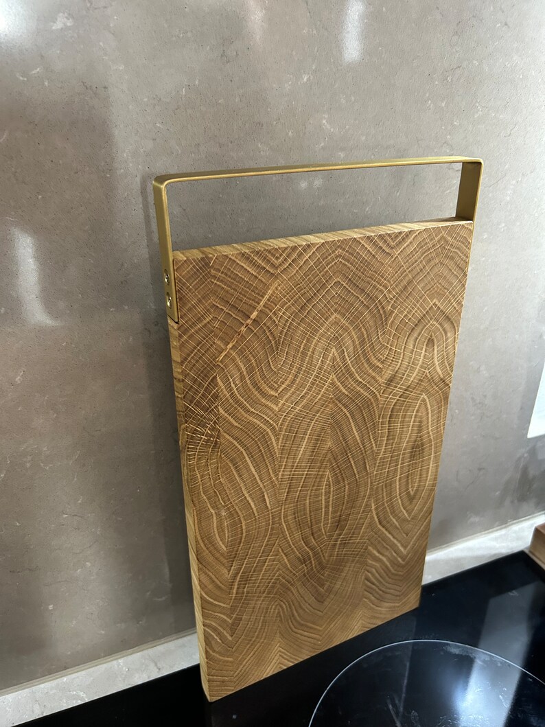 May include: A wooden cutting board with a gold metal handle. The board has a natural wood grain pattern and is rectangular in shape.