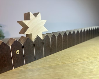 Puede incluir: Un calendario de adviento de madera con una tapa en forma de estrella. El calendario tiene 24 pequeñas puertas, cada una con un número. La estrella está encima de la puerta con el número 6.
