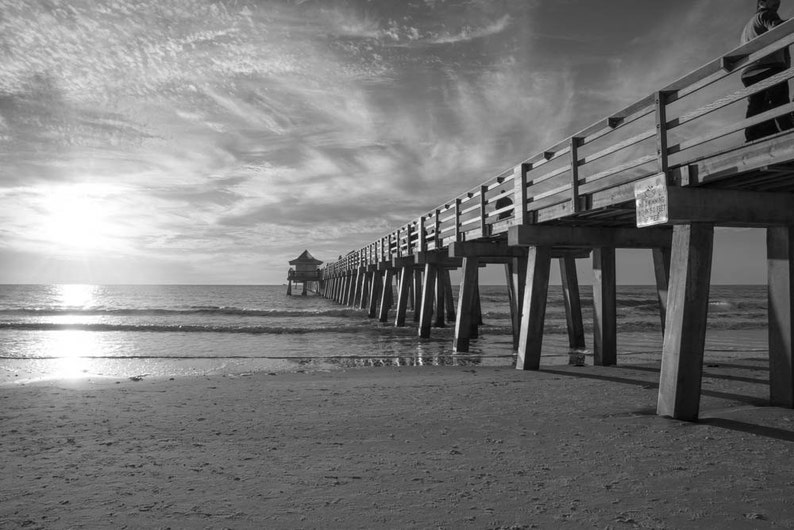 Puede incluir: Una foto en blanco y negro de un muelle de madera que se extiende sobre un mar tranquilo. El muelle tiene una barandilla de madera y est&aacute; sostenido por pilares. El cielo est&aacute; nublado y el sol brilla.