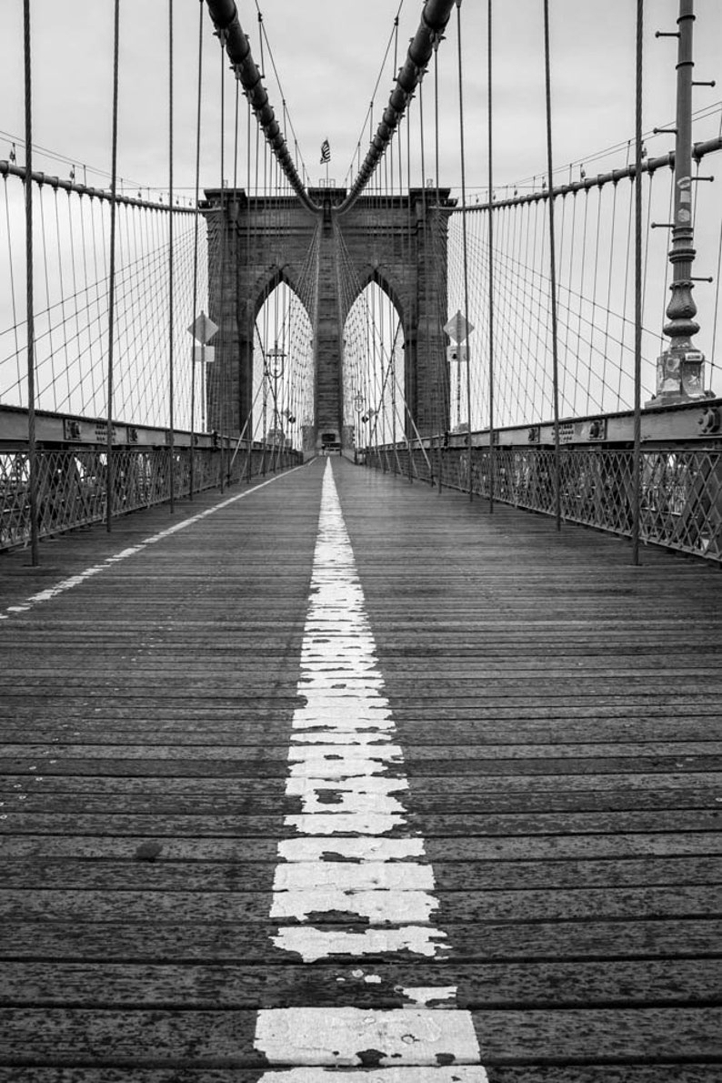 Puede incluir: Una fotograf&iacute;a en blanco y negro del puente de Brooklyn en la ciudad de Nueva York. La foto muestra la pasarela del puente con una l&iacute;nea blanca pintada en el centro. Las torres del puente y los cables son visibles en el fondo.
