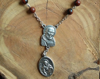 Op de afbeelding: Een bruin en zwart gebedssnoer met een zilveren hanger met een afbeelding van Sint Padre Pio. De hanger is bevestigd aan een zilveren ketting.