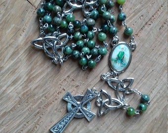 May include: A green stone rosary with a silver Celtic cross and a silver charm with a picture of a saint. The rosary is on a wooden surface.