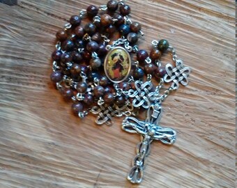 Op de afbeelding: Een rozenkrans met bruine kralen en een zilveren kruisbeeld. De kralen zijn een mix van bruine en bruinachtige kleuren. Het kruisbeeld en de verbindingsketting zijn zilver. Een ovale hanger met een religieuze afbeelding is ook aanwezig.
