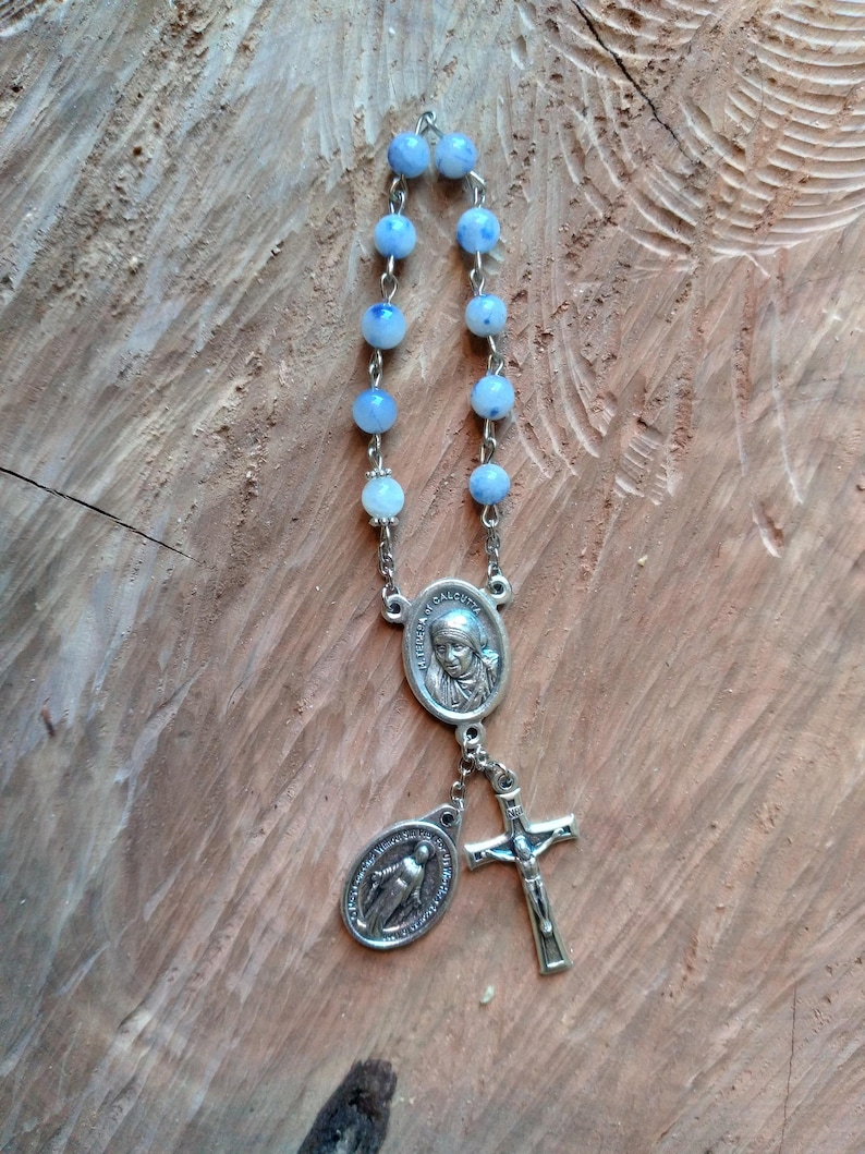Op de afbeelding: Een zilveren rozenkrans met blauwe kralen en een zilveren hanger met een afbeelding van Moeder Teresa. De hanger is bevestigd aan een zilveren kruis.
