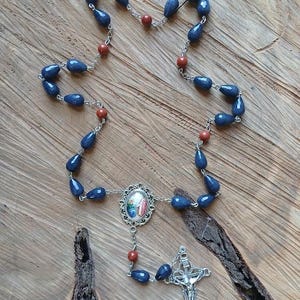 Op de afbeelding: Een rozenkrans met traanvormige donkerblauwe kralen en ronde roodbruine kralen verbonden door zilverkleurige metalen schakels. Het middelpunt is een ovaal metalen frame met een kleine religieuze afbeelding en een zilverkleurig kruisbeeld.