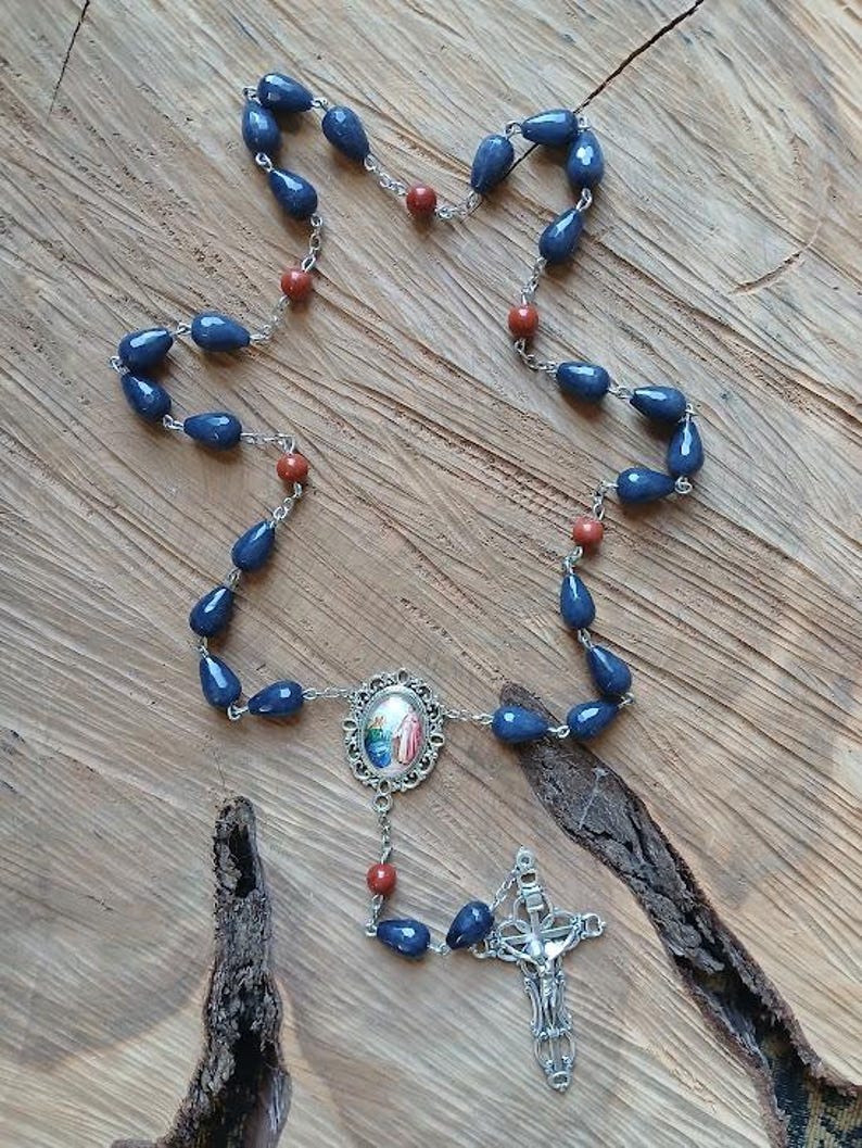 Op de afbeelding: Een rozenkrans met traanvormige donkerblauwe kralen en ronde roodbruine kralen verbonden door zilverkleurige metalen schakels. Het middelpunt is een ovaal metalen frame met een kleine religieuze afbeelding en een zilverkleurig kruisbeeld.