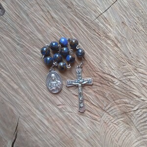 May include: A rosary with blue and brown speckled beads, a silver crucifix, and a silver pendant with a religious figure. The rosary is displayed on a textured wooden surface.