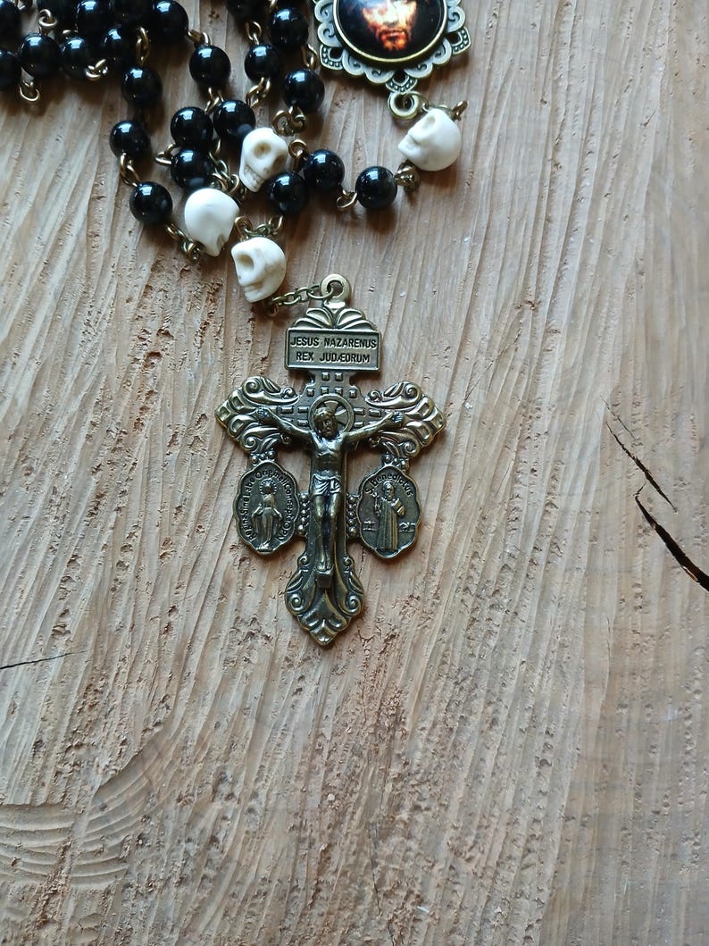 Op de afbeelding: Een rozenkrans met zwarte kralen en schedelvormige kralen. Het kruisbeeld is bronskleurig met de inscriptie "JESUS NAZARENUS REX JUDAEORUM". De rozenkrans ligt op een houten oppervlak.