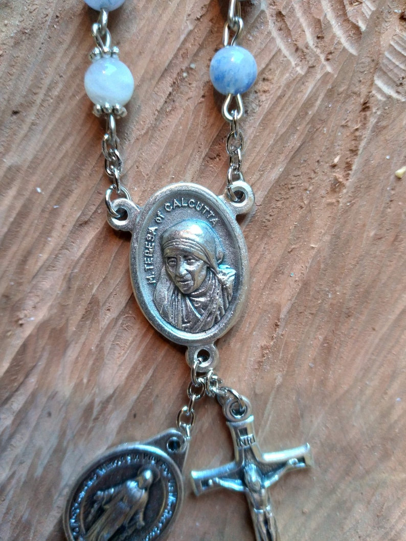 Op de afbeelding: Een zilveren rozenkrans met blauwe en witte kralen. Het middelpunt is een zilveren ovaal medaillon met een afbeelding van Moeder Teresa van Calcutta. De rozenkrans bevat ook een zilveren kruisbeeld en een zilveren ovaal medaillon met een afbeelding van de Maagd Maria.