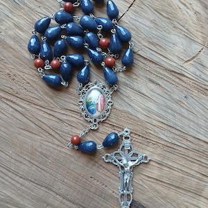 Op de afbeelding: Een rozenkrans met traanvormige donkerblauwe kralen en ronde roodbruine kralen verbonden door zilverkleurige metalen schakels. Het middelpunt is een ovale medaille met een religieuze afbeelding en het kruisbeeld is een sierlijk zilverkleurig kruis.