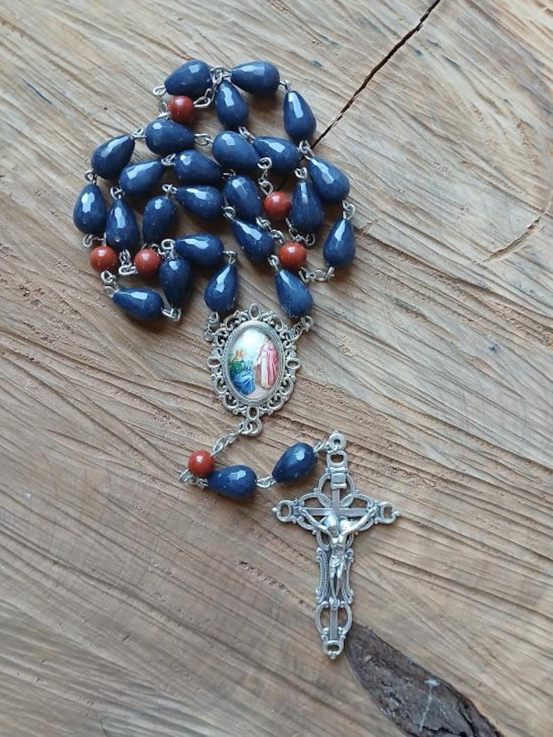 Op de afbeelding: Een rozenkrans met traanvormige donkerblauwe kralen en ronde roodbruine kralen verbonden door zilverkleurige metalen schakels. Het middelpunt is een ovale medaille met een religieuze afbeelding en het kruisbeeld is een sierlijk zilverkleurig kruis.