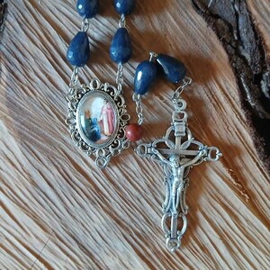 Op de afbeelding: Een rozenkrans met donkerblauwe, gefacetteerde, druppelvormige kralen verbonden door zilverkleurige metalen schakels. Het bevat een ovaalvormig middenstuk met een religieuze afbeelding en een zilverkleurig kruisbeeld met een Jezusfiguur.
