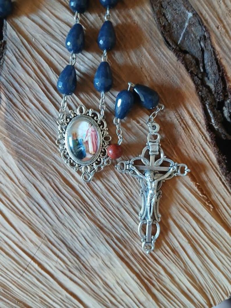 Op de afbeelding: Een rozenkrans met donkerblauwe, gefacetteerde, druppelvormige kralen verbonden door zilverkleurige metalen schakels. Het bevat een ovaalvormig middenstuk met een religieuze afbeelding en een zilverkleurig kruisbeeld met een Jezusfiguur.