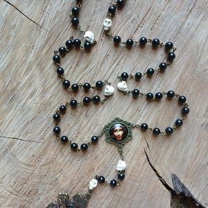 Op de afbeelding: Een zwarte kralenrozenkrans met schedelaccenten en een kruisbeeld in bronskleur. De rozenkrans heeft een centrale hanger met een religieus beeld, gepresenteerd op een houten oppervlak.