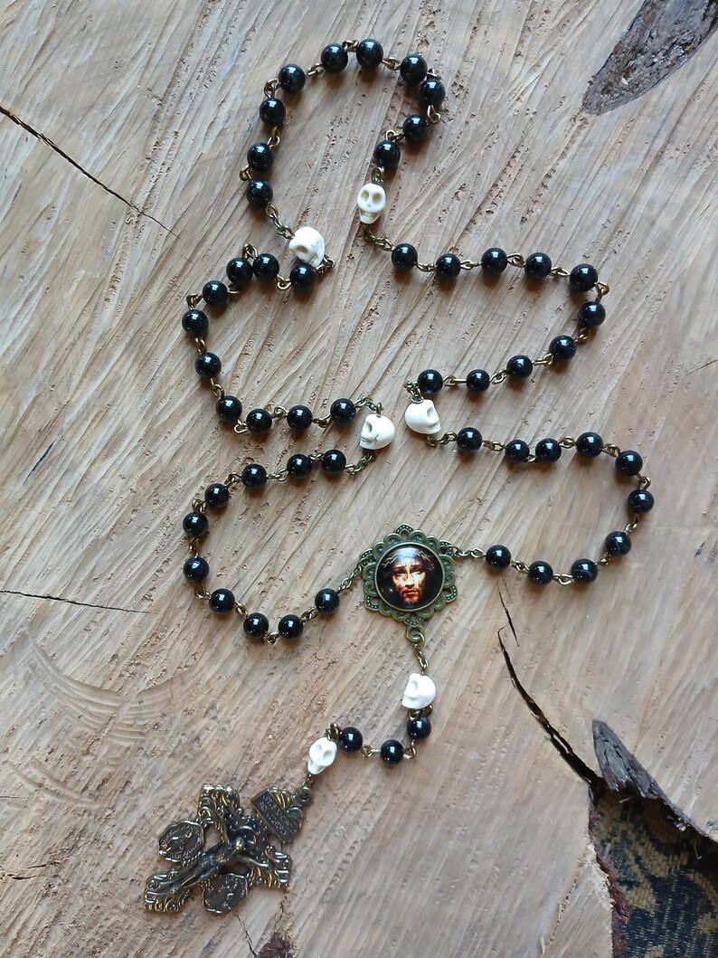 Op de afbeelding: Een zwarte kralenrozenkrans met schedelaccenten en een kruisbeeld in bronskleur. De rozenkrans heeft een centrale hanger met een religieus beeld, gepresenteerd op een houten oppervlak.