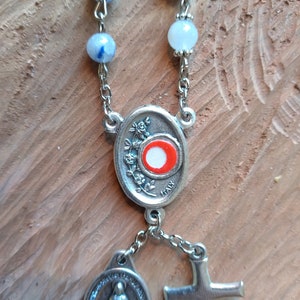 Op de afbeelding: Een zilveren rozenkrans met blauwe en witte kralen, een zilveren ovale bedel met een rood en wit centrum, een zilveren bedel met een afbeelding van de Maagd Maria en een zilveren kruis.