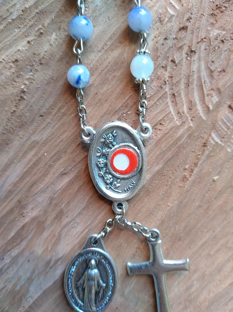 Op de afbeelding: Een zilveren rozenkrans met blauwe en witte kralen, een zilveren ovale bedel met een rood en wit centrum, een zilveren bedel met een afbeelding van de Maagd Maria en een zilveren kruis.