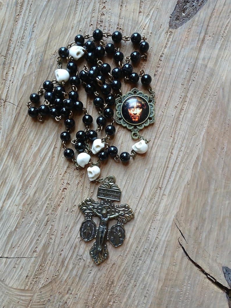 Op de afbeelding: Een rozenkrans met zwarte kralen en schedelvormige kralen. Het heeft een bronzen kruisbeeldhanger en een ronde hanger met een afbeelding van Jezus. De rozenkrans wordt gepresenteerd op een houten oppervlak.