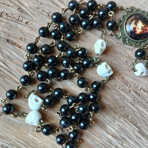 Op de afbeelding: Een rozenkrans met zwarte kralen en kleine schedelaccenten. De rozenkrans heeft een decoratieve hanger met een afbeelding en een metalen kruis. De kralen zijn geregen aan een goudkleurige ketting.