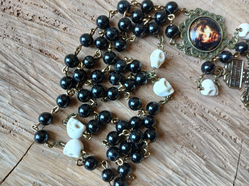 Op de afbeelding: Een rozenkrans met zwarte kralen en kleine schedelaccenten. De rozenkrans heeft een decoratieve hanger met een afbeelding en een metalen kruis. De kralen zijn geregen aan een goudkleurige ketting.