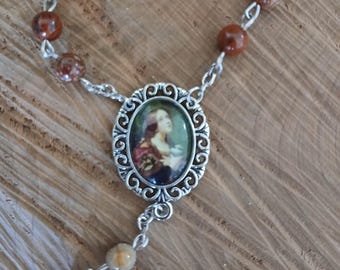 Op de afbeelding: Een rozenkrans met bruine en bruinbeige stenen kralen en zilverkleurige metalen schakels. Het middelpunt heeft een ovale hanger met een religieuze afbeelding. De kralen zijn verbonden door fijne zilveren kettingen, waardoor een traditioneel religieus accessoire ontstaat.