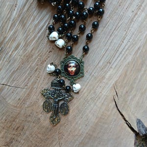 Op de afbeelding: Een rozenkransketting met zwarte kralen en schedelaccenten. De ketting bevat een kruisbeeldhanger en een ronde hanger met een religieuze afbeelding. De metalen elementen hebben een antieke messing afwerking.