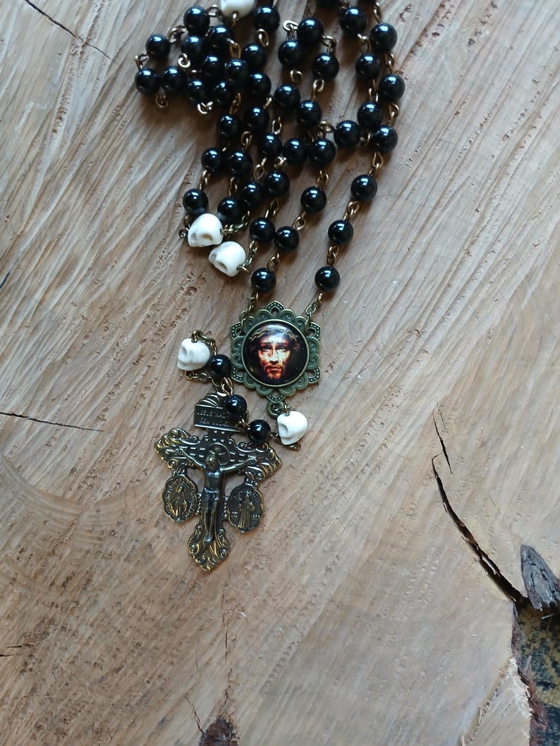 Op de afbeelding: Een rozenkransketting met zwarte kralen en schedelaccenten. De ketting bevat een kruisbeeldhanger en een ronde hanger met een religieuze afbeelding. De metalen elementen hebben een antieke messing afwerking.