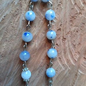 Op de afbeelding: Een zilveren ketting met blauwe en witte kralen. De kralen hebben een gevlekt patroon. De ketting heeft een zilveren hanger aan het einde.