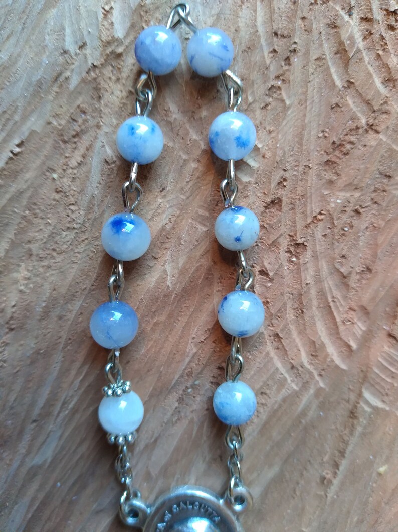 Op de afbeelding: Een zilveren ketting met blauwe en witte kralen. De kralen hebben een gevlekt patroon. De ketting heeft een zilveren hanger aan het einde.