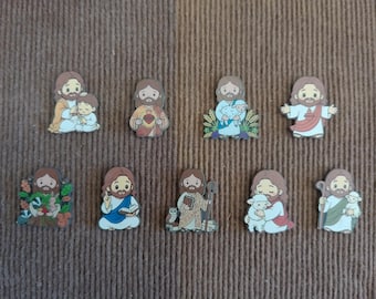 Op de afbeelding: Een verzameling van negen kleine, platte pins in cartoonstijl met verschillende afbeeldingen van Jezus. Elke pin toont een andere scène, waaronder Jezus met een kind, een hart, een lam, of omringd door bosdieren en natuurlijke elementen.