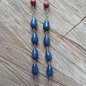 Op de afbeelding: Een ketting met twee parallelle zilverkleurige metalen kettingen, versierd met afwisselend donkerblauwe druppelvormige kralen en kleinere ronde roodbruine kralen. De kralen zijn verticaal met elkaar verbonden door kleine metalen ringetjes.