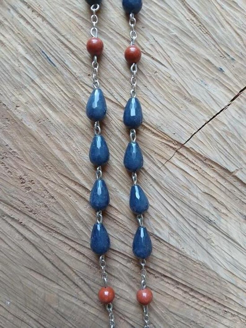 Op de afbeelding: Een ketting met twee parallelle zilverkleurige metalen kettingen, versierd met afwisselend donkerblauwe druppelvormige kralen en kleinere ronde roodbruine kralen. De kralen zijn verticaal met elkaar verbonden door kleine metalen ringetjes.