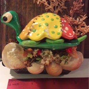 Pode incluir: Uma figura de tartaruga caprichosa com cabeça e carapaça verdes, carapaça amarela e vermelha e uma base adornada com conchas e flores secas. Uma figura de joaninha fica em cima da tartaruga. A figura tem aproximadamente 10 cm de comprimento.
