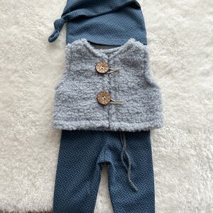 K&ouml;nnte beinhalten: Ein Baby-Gnom-Outfit mit einer grauen Weste, einer blauen Hose und einer blauen M&uuml;tze. Die Weste hat Holzkn&ouml;pfe.