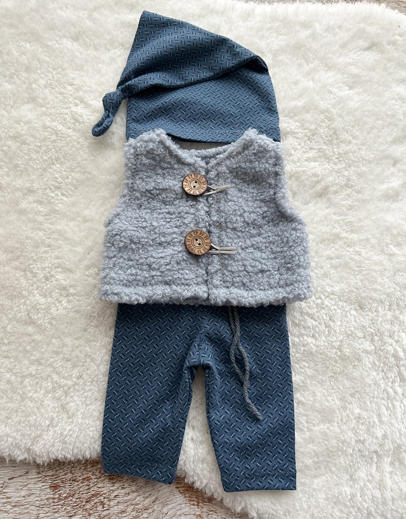 K&ouml;nnte beinhalten: Ein Baby-Gnom-Outfit mit einer grauen Weste, einer blauen Hose und einer blauen M&uuml;tze. Die Weste hat Holzkn&ouml;pfe.