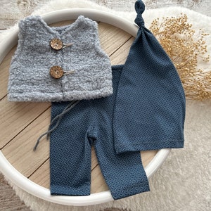 K&ouml;nnte beinhalten: Ein Baby-Outfit-Set mit einer grauen, flauschigen Weste, einer blauen Hose und einer blauen Strickm&uuml;tze. Die Weste hat zwei Holzkn&ouml;pfe.