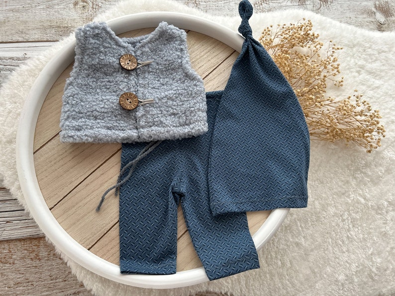 K&ouml;nnte beinhalten: Ein Baby-Outfit-Set mit einer grauen, flauschigen Weste, einer blauen Hose und einer blauen Strickm&uuml;tze. Die Weste hat zwei Holzkn&ouml;pfe.