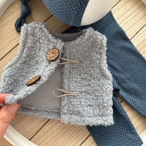 K&ouml;nnte beinhalten: Eine graue, flauschige Weste mit zwei Holzkn&ouml;pfen und einem darunter liegenden Stoff mit blau-wei&szlig;em Muster.