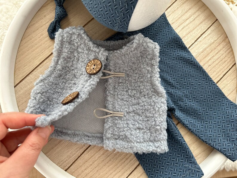 K&ouml;nnte beinhalten: Eine graue, flauschige Weste mit zwei Holzkn&ouml;pfen und einem darunter liegenden Stoff mit blau-wei&szlig;em Muster.