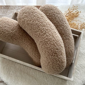 May include: Two beige, fluffy, crescent-shaped pillows in a wooden tray. The pillows are made of a soft, fuzzy material.