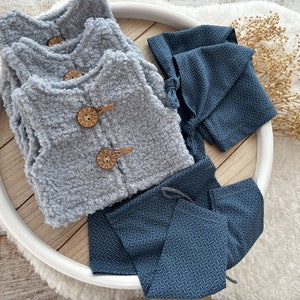 K&ouml;nnte beinhalten: Ein Set aus drei grauen, flauschigen Westen mit Holzkn&ouml;pfen und zwei blauen, gemusterten Stoffteilen, eines mit einer spitzen Kapuze und das andere mit einem Kordelzug, auf einem Holztablett.