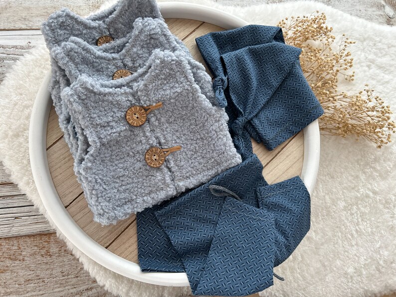 K&ouml;nnte beinhalten: Ein Set aus drei grauen, flauschigen Westen mit Holzkn&ouml;pfen und zwei blauen, gemusterten Stoffteilen, eines mit einer spitzen Kapuze und das andere mit einem Kordelzug, auf einem Holztablett.