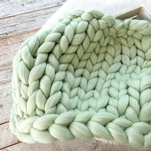 Peut inclure: Une couverture en tricot épais de couleur vert clair, réalisée avec un fil épais. La couverture est pliée en deux et a une texture douce et duveteuse.