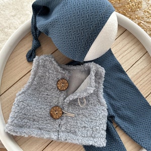 K&ouml;nnte beinhalten: Eine graue, flauschige Weste mit zwei Holzkn&ouml;pfen und einer passenden blauen und wei&szlig;en gemusterten M&uuml;tze und Hose.