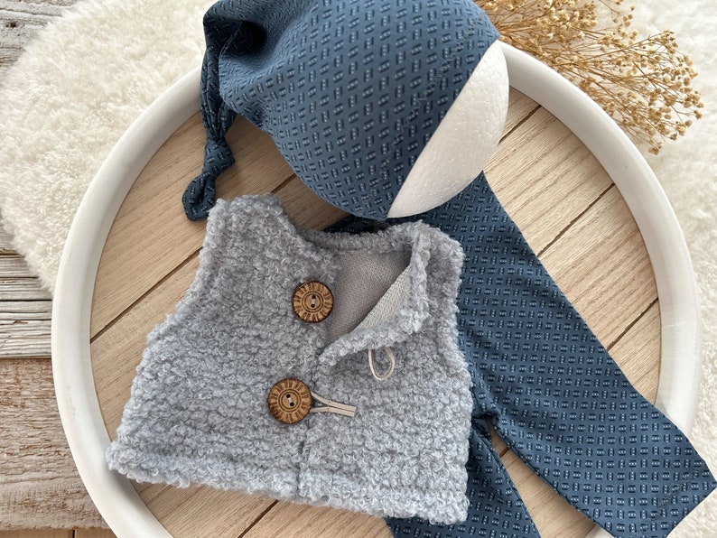 K&ouml;nnte beinhalten: Eine graue, flauschige Weste mit zwei Holzkn&ouml;pfen und einer passenden blauen und wei&szlig;en gemusterten M&uuml;tze und Hose.